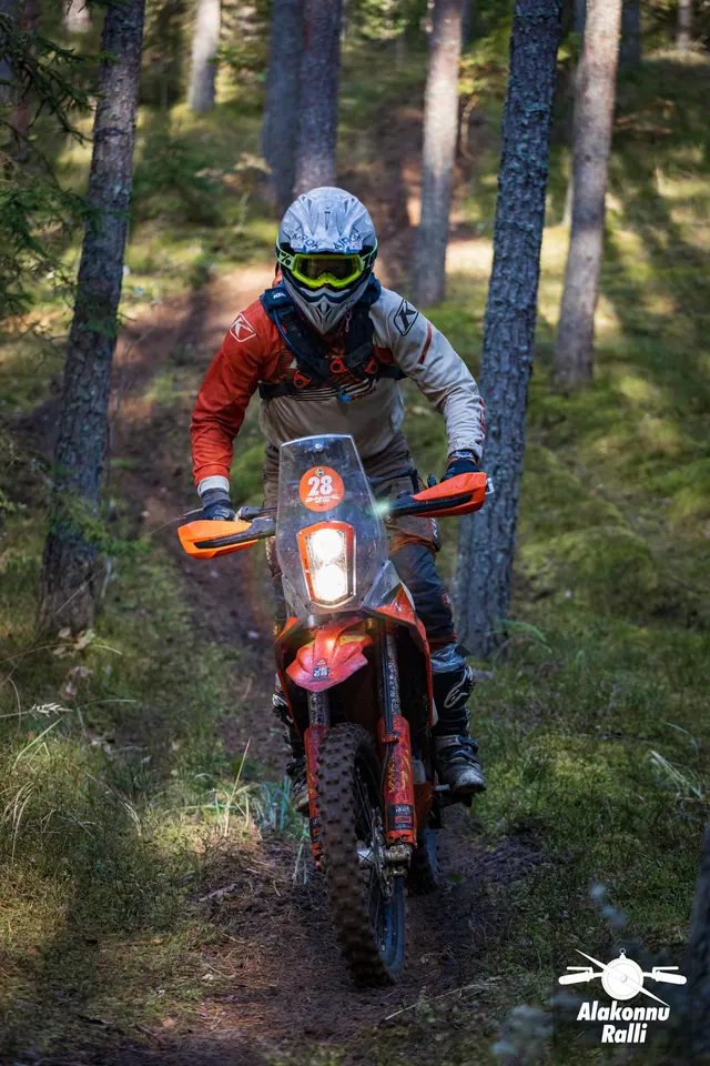Alakonnu Ralli 2026: oranži ja halli sõiduvarustusega mootorrattur läheneb kõrge tuuleklaasiga rattal kitsal männimetsarajal.