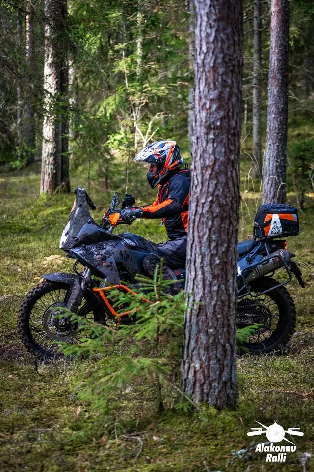 Alakonnu Ralli 2026: mootorrattur oranži raamiga rattal osaliselt männipuu taga.