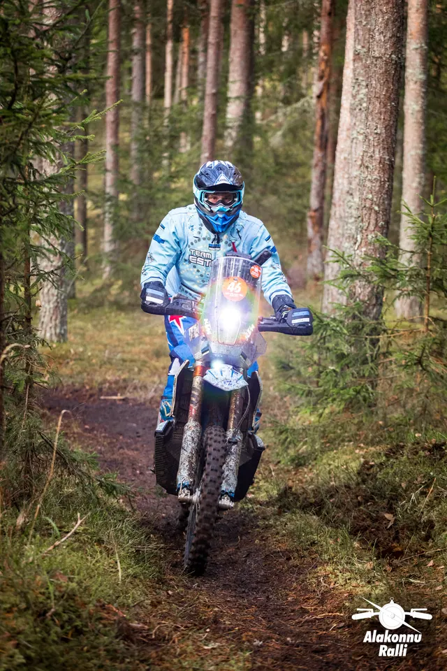 Alakonnu Ralli 2026: heledas sinises varustuses sõitja läheneb ereda esitulega metsarajal.