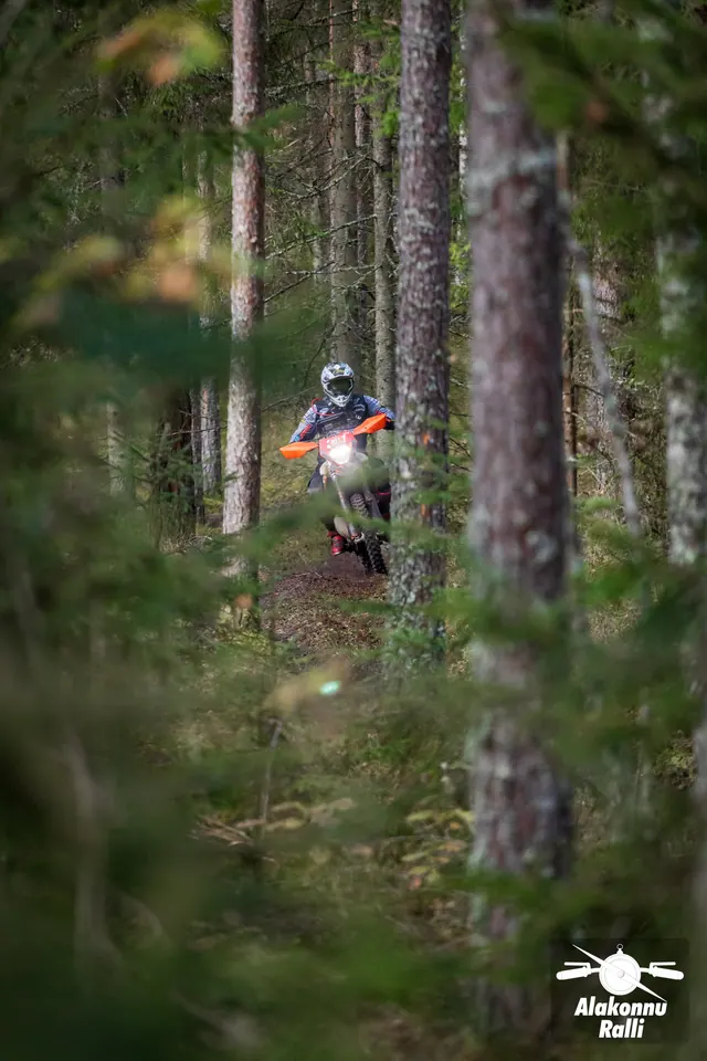Alakonnu Ralli 2026: mootorrattur oranžide käekaitsmetega rattal paistab puude tagant.