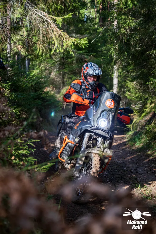 Alakonnu Ralli 2026: oranžis varustuses sõitja kallutab suurt matkaenduromootorratast metsarajal.