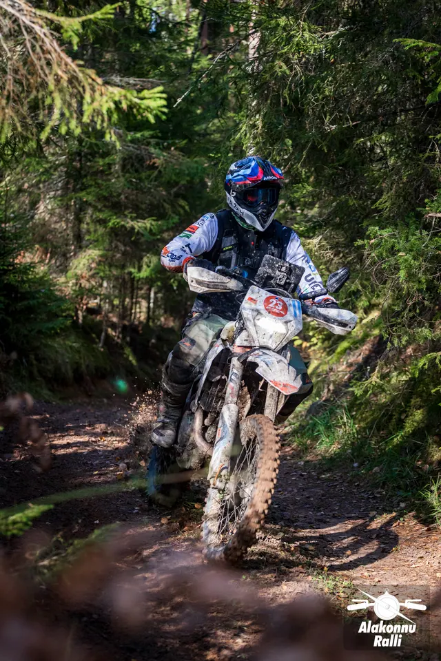 Alakonnu Rally 2026: a rider on a white motorcycle approaches at a bend lined with spruce trees.