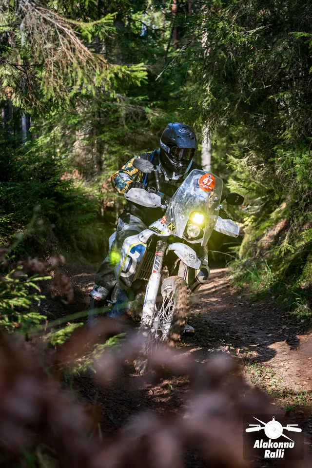Alakonnu Rally 2026: a motorcyclist with a yellow headlight rides on a forest trail dappled with sunlight.