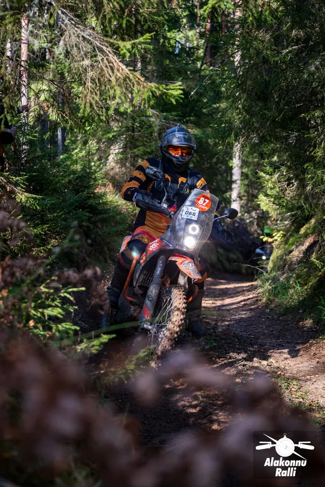 Alakonnu Rally 2026: a motorcyclist in orange and black gear at a narrow bend between spruce trees.