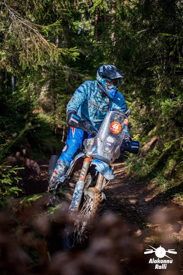 Alakonnu Rally 2026: a rider in blue gear on a bike with a tall windscreen at a forest bend.