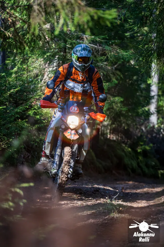Alakonnu Rally 2026: a rider in orange gear on a bike with its headlight on at a forest bend.