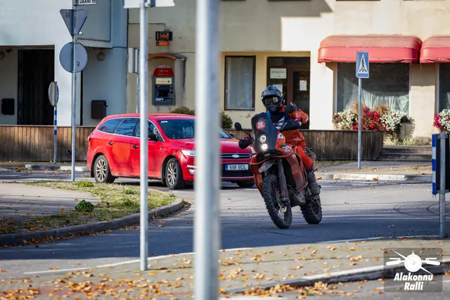 Alakonnu Ralli 2026: oranžil mootorrattal sõitja läbib tänavakurvi hoonete ja punase auto ees.