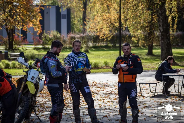 Alakonnu Ralli 2026: kolm sõiduvarustuses mootorratturit seisavad pargitud ratta kõrval; kahel on toidunõu käes ja üks vaatab telefoni.