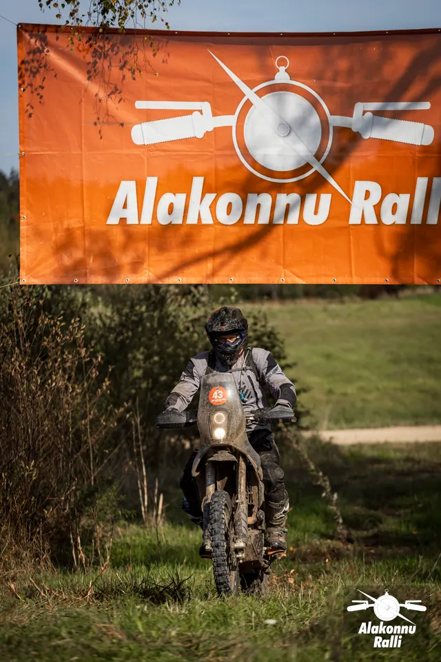 Alakonnu Ralli 2026: halli varustusega mootorrattur rohurajal oranži Alakonnu Ralli bänneri all.