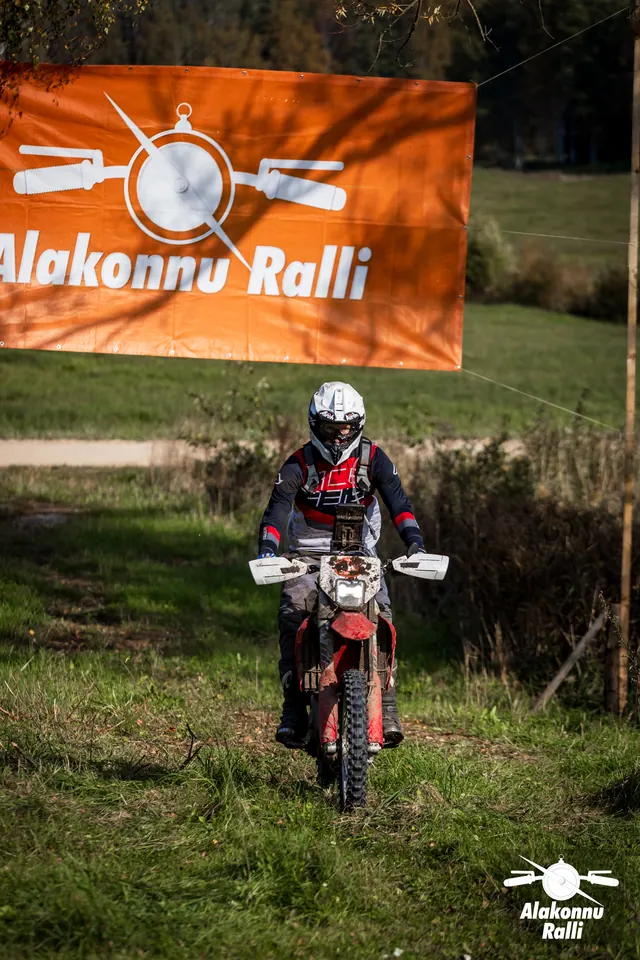 Alakonnu Ralli 2026: valge kiivriga sõitja punasel rattal läheneb oranži rallibänneri alt.