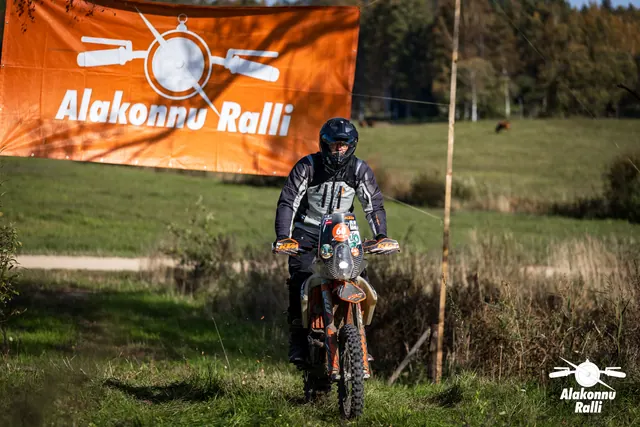 Alakonnu Ralli 2026: tumedas varustuses sõitja oranžil rattal rohurajal rallibänneri ees.