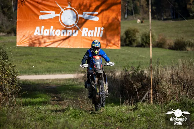 Alakonnu Ralli 2026: sinises varustuses sõitja läheneb rohurajal oranži Alakonnu Ralli bänneri alt.