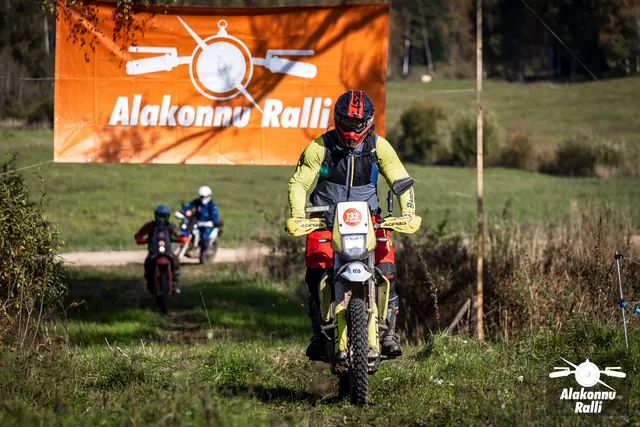 Alakonnu Ralli 2026: kollase jakiga sõitja läheneb rohurajal rallibänneri alt, teised taga.