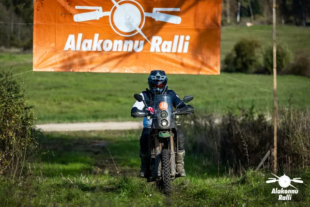 Alakonnu Ralli 2026: sinise ja valge varustusega mootorrattur läheneb rohurajal rallibänneri alt.