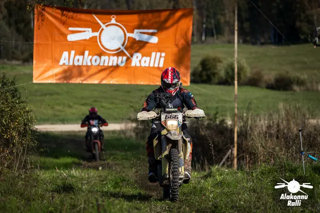 Alakonnu Ralli 2026: punase kiivriga sõitja läheneb mööda rohurada, taustal teine mootorratas ja oranž rallibänner.