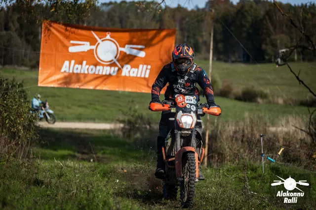 Alakonnu Ralli 2026: sõitja läheneb oranžide käekaitsmetega mootorrattal mööda rohurada rallibänneri all.