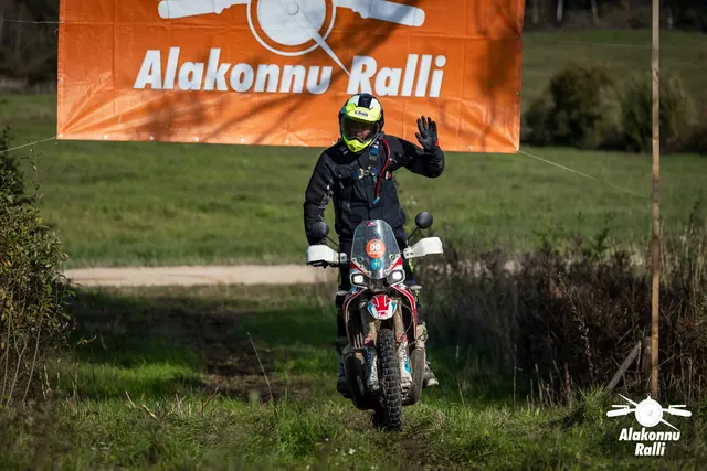 Alakonnu Ralli 2026: neoonkollase kiivriga sõitja lehvitab mootorrattalt oranži rallibänneri all.