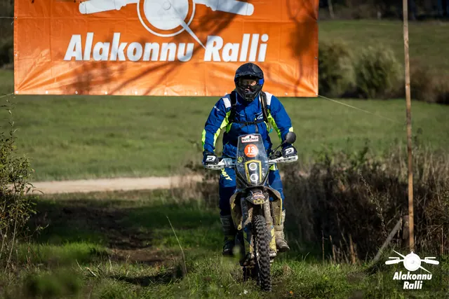 Alakonnu Ralli 2026: sinises ja kollases sõiduvarustuses osaleja sõidab mööda rohurada rallibänneri all.