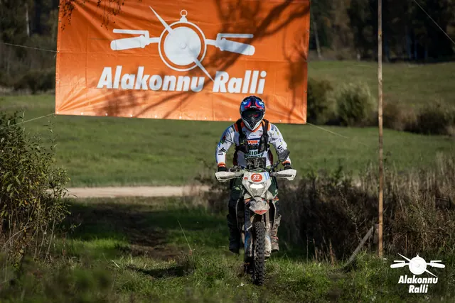 Alakonnu Ralli 2026: valges ja hallis sõiduvarustuses osaleja läheneb mööda rohurada oranži rallibänneri all.
