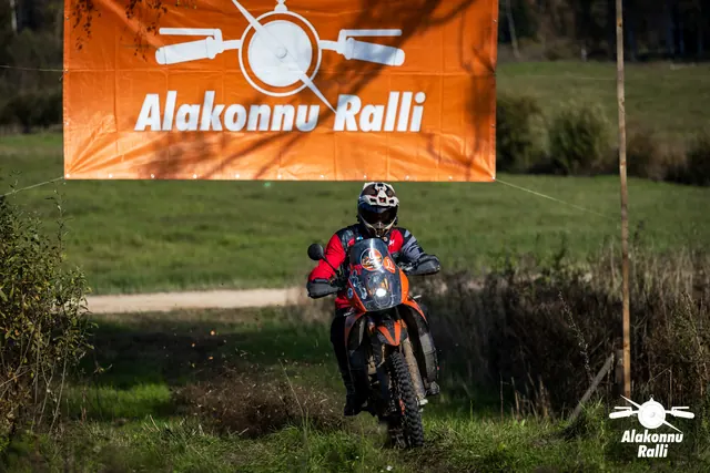 Alakonnu Ralli 2026: punase ja tumeda sõiduvarustusega osaleja läheneb oranžil mootorrattal mööda rohurada.
