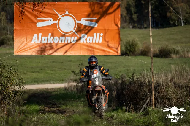 Alakonnu Ralli 2026: tumedas sõiduvarustuses osaleja läheneb oranžil mootorrattal rallibänneri all.