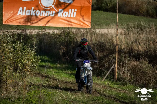Alakonnu Ralli 2026: tumedas sõiduvarustuses osaleja sõidab mööda rohurada, oranž rallibänner taustal.