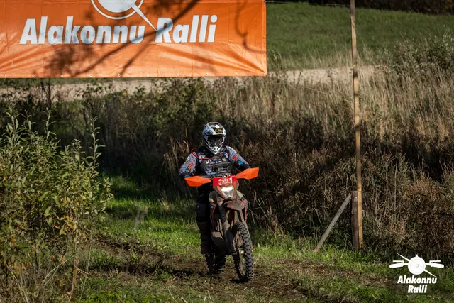 Alakonnu Ralli 2026: sõitja läheneb oranžide käekaitsmetega mootorrattal mööda rohurada rallibänneri all.