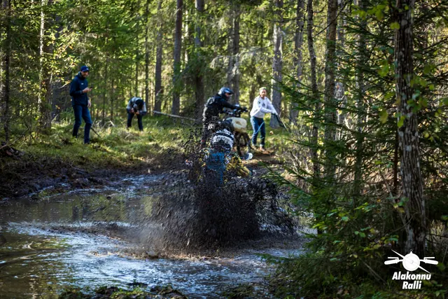 Alakonnu Ralli 2026: mootorrattur paiskab metsaraja veetakistuses mudast vett üles, kaldal jälgivad inimesed.
