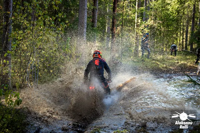 Alakonnu Rally 2026: a motorcyclist rides through a flooded forest track and throws a high spray behind.