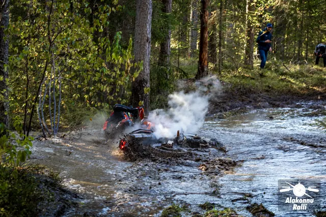 Alakonnu Rally 2026: a rider is beside a motorcycle lying on its side in a flooded track, surrounded by splashing water and a pale cloud.