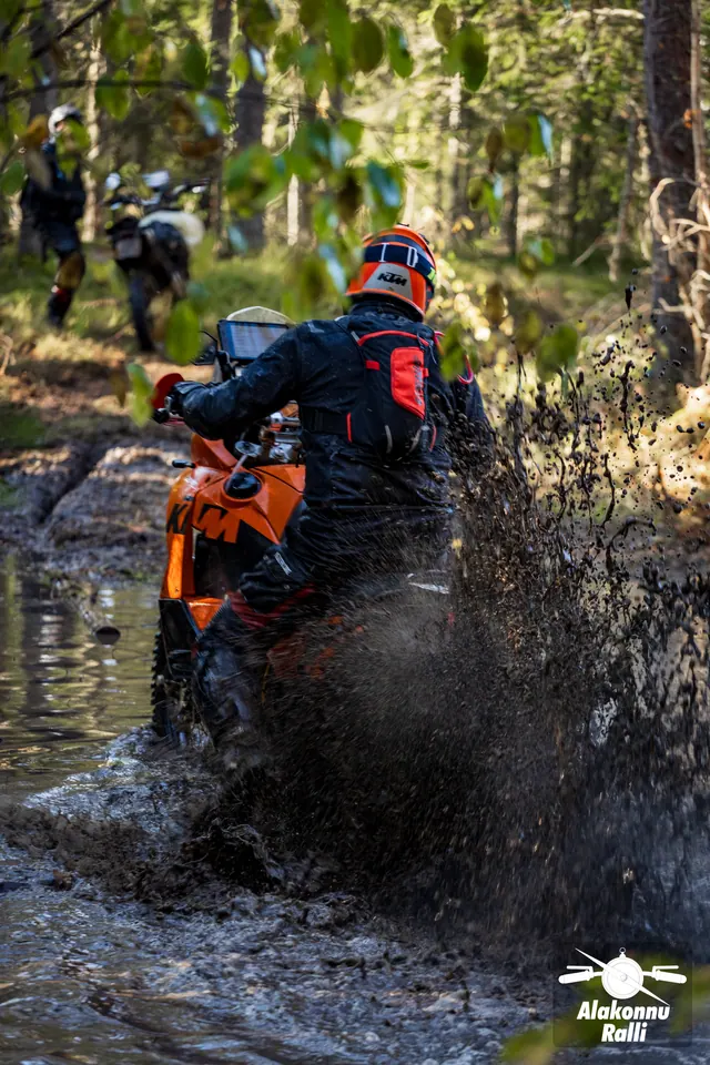 Alakonnu Rally 2026: a rider on an orange motorcycle throws up muddy water in a flooded forest track.