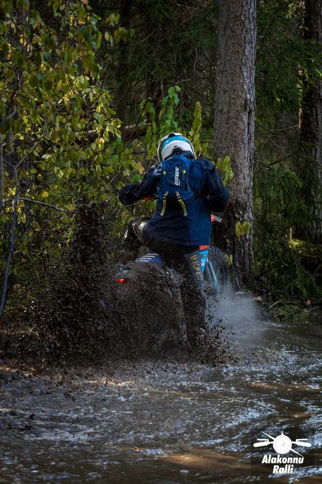 Alakonnu Rally 2026: a rider in a blue helmet moves through a flooded forest track with water spraying behind the rear wheel.