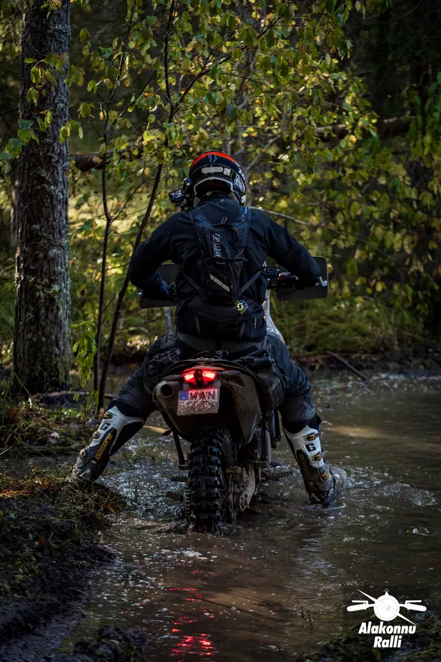 Alakonnu Rally 2026: a motorcyclist puts both feet down in the flooded track and moves between the trees.