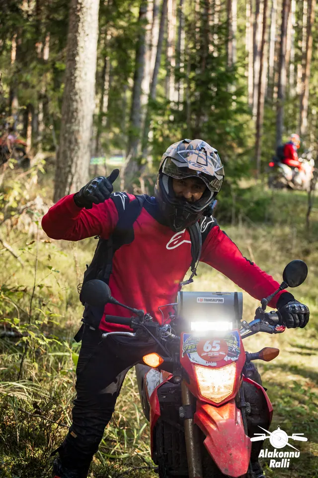 Alakonnu Rally 2026: a participant in a red riding jersey waves beside an orange motorcycle at the edge of the forest.