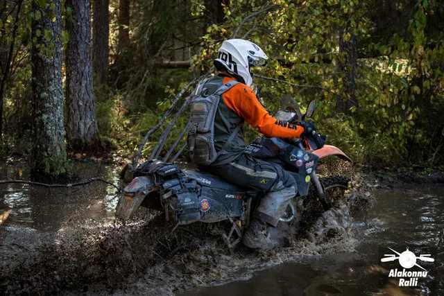 Alakonnu Rally 2026: a rider in a white helmet crosses muddy water on a forest track on an orange motorcycle.