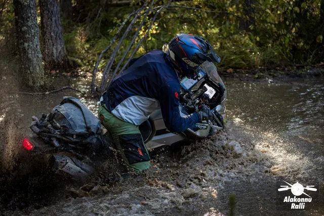 Alakonnu Rally 2026: a participant in a blue top holds a motorcycle leaning heavily to one side in the flooded track.