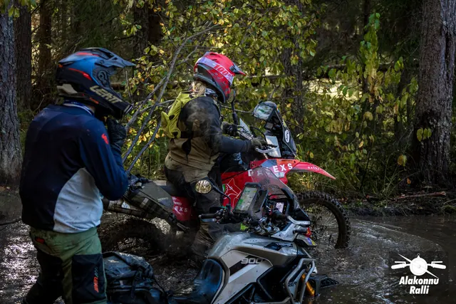 Alakonnu Rally 2026: a rider in a red helmet moves through the flooded track on a red motorcycle with another participant standing nearby.