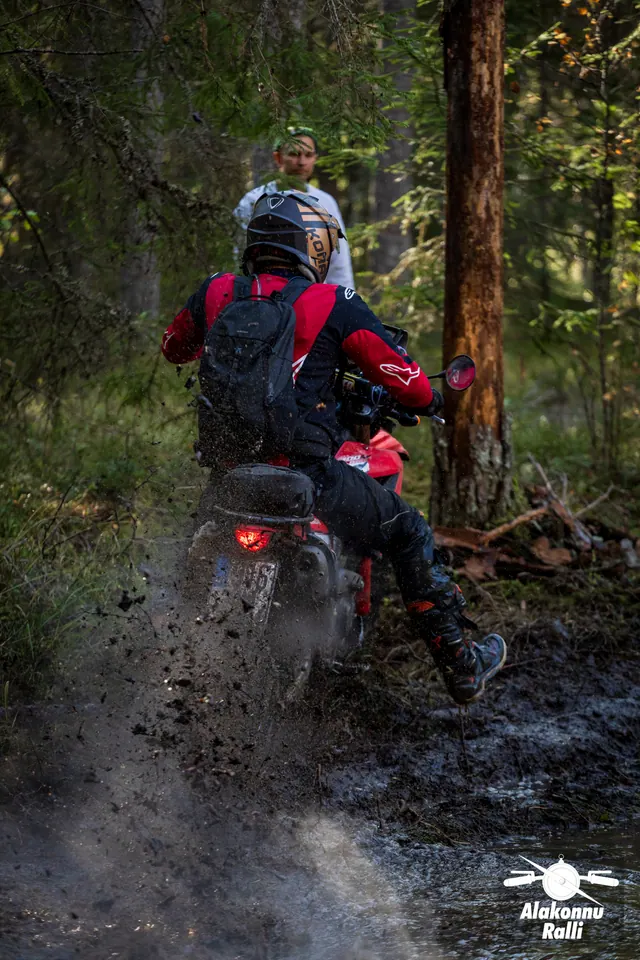 Alakonnu Rally 2026: a participant in a red riding jersey rides out of the flooded track into the forest with water spraying behind.