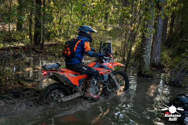Alakonnu Rally 2026: a rider with an orange backpack moves through water on a forest track on an orange motorcycle.