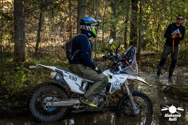 Alakonnu Rally 2026: a motorcyclist puts both feet down on a white motorcycle in the flooded track while a person stands on the bank.