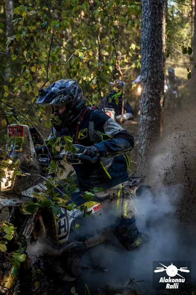 Alakonnu Rally 2026: a rider crosses muddy water beside a tree with spray and a pale cloud rising around the motorcycle.
