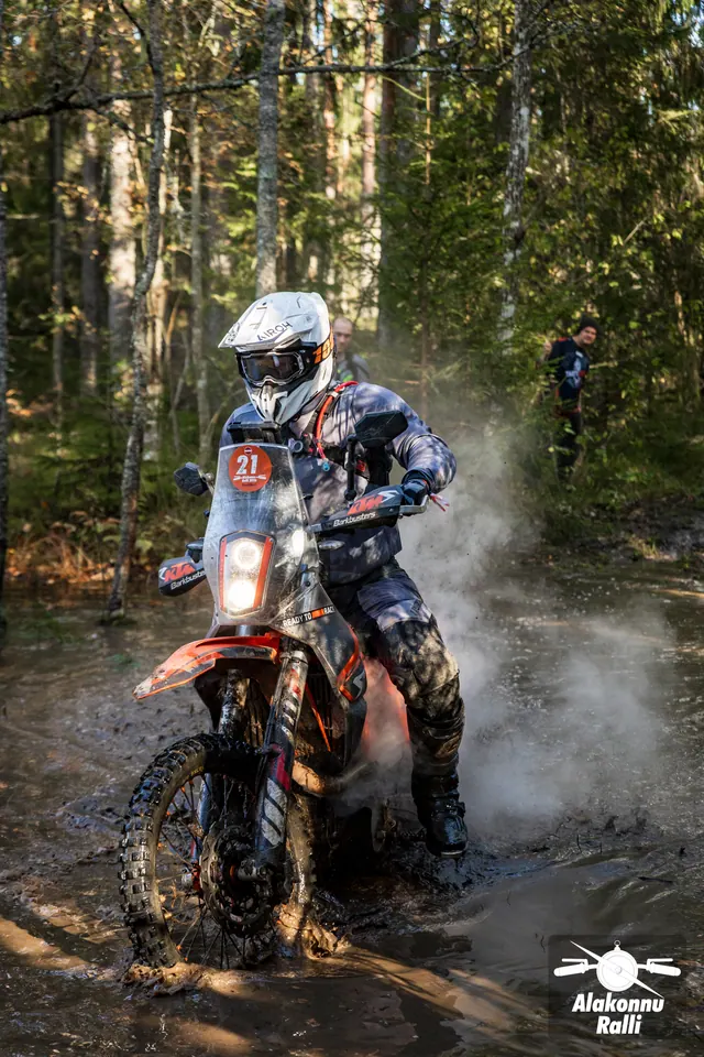 Alakonnu Rally 2026: a rider in a white helmet approaches through water on a forest track on an orange motorcycle.