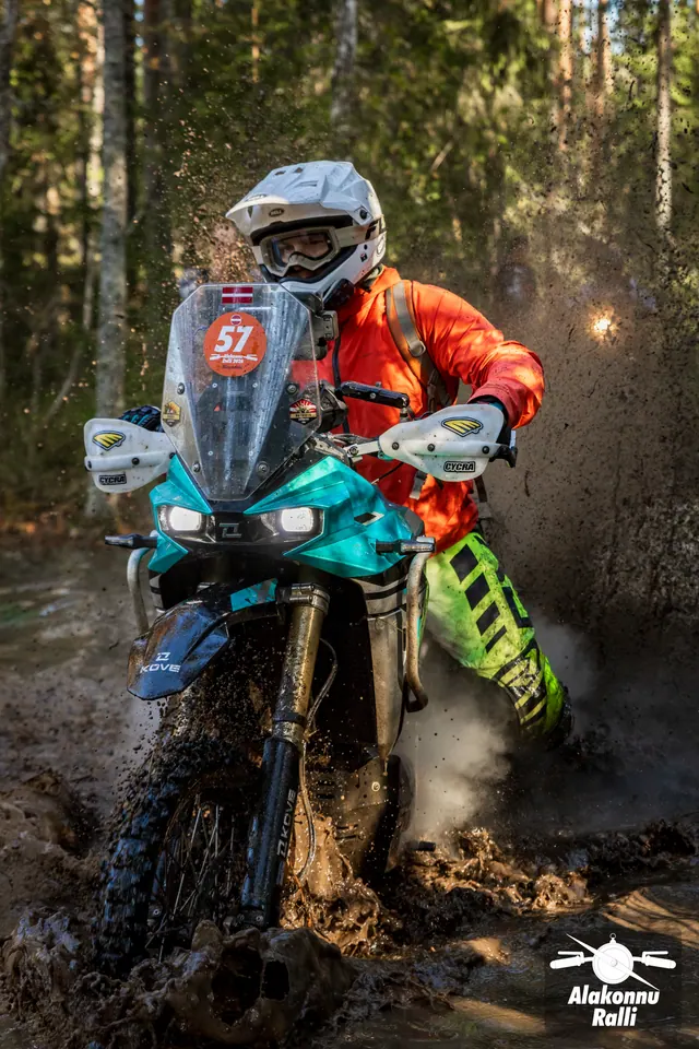 Alakonnu Rally 2026: a participant in orange riding gear and a white helmet approaches through muddy water on a green motorcycle.
