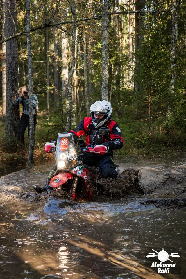 Alakonnu Rally 2026: a participant in a white helmet and red riding gear approaches through water on a forest track.