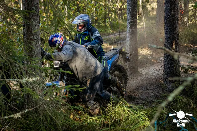 Alakonnu Ralli 2026: inimene lükkab mootorratta esiosa puude vahel, kiivriga sõitja hoiab ratast tagant.