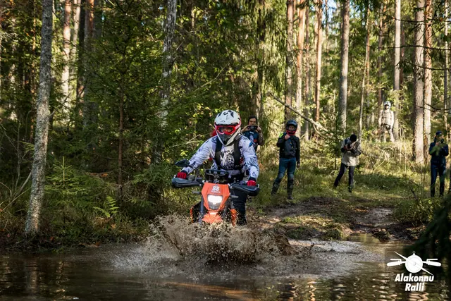 Alakonnu Ralli 2026: valge kiivriga sõitja siseneb oranžil mootorrattal metsaraja veetakistusse, taustal jälgivad inimesed.