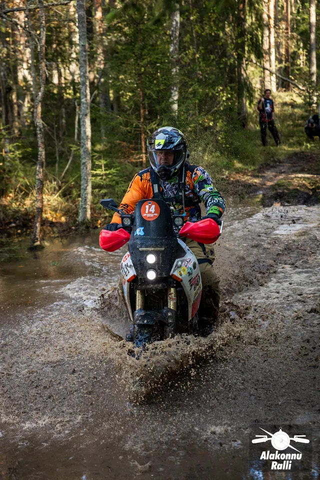 Alakonnu Ralli 2026: sõitja läheneb punaste käekaitsmetega mootorrattal läbi metsaraja vee.