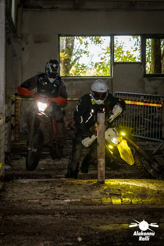 Alakonnu Ralli 2026: kaks mootorratturit läheneb hoone kitsas rajakoridoris põlevate esituledega.