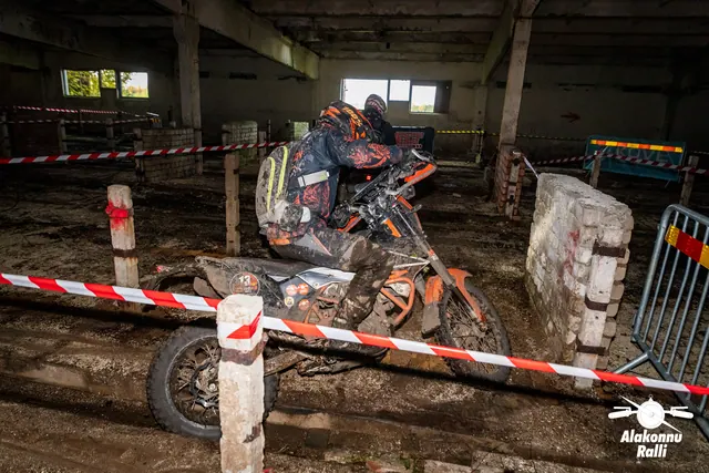 Alakonnu Ralli 2026: tumedas ja oranžis sõiduvarustuses osaleja juhib oranži mootorratast hoones müüri kõrvalt.