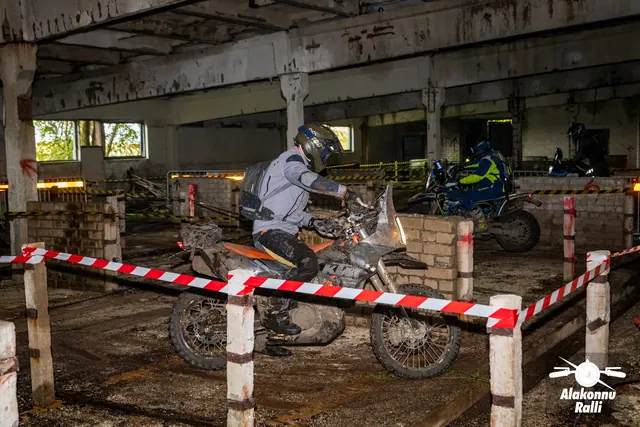 Alakonnu Ralli 2026: hallis sõiduvarustuses osaleja pöörab mootorrattaga hoones müüri kõrval, teine sõitja taustal.